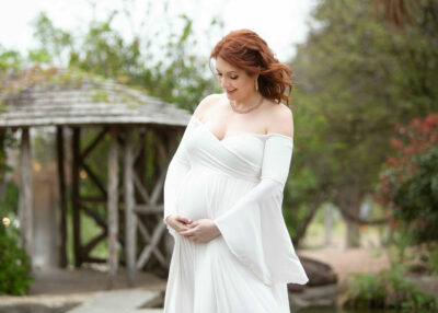 A pregnant woman in a white dress poses in front of a pond.