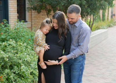 A pregnant woman and her husband are holding their baby girl in front of a brick wall.