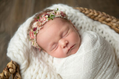 A newborn girl in a basket wearing a flower headband.