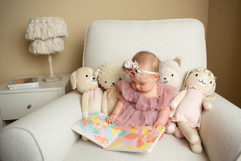 A baby girl is sitting in a chair reading a book with stuffed animals.