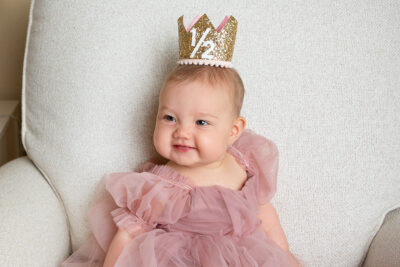 A baby girl in a pink dress with a gold crown sitting on a chair.