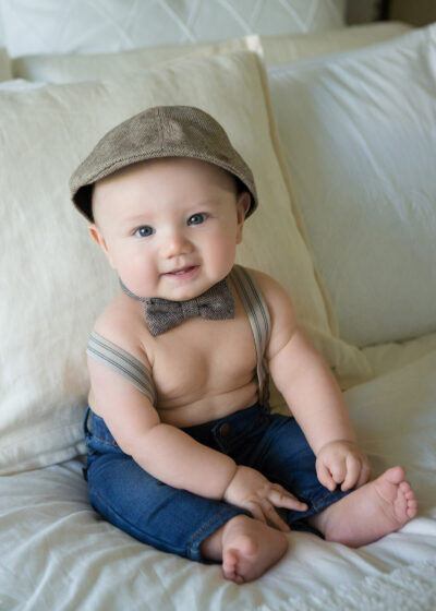 A baby wearing a hat and suspenders sitting on a bed.