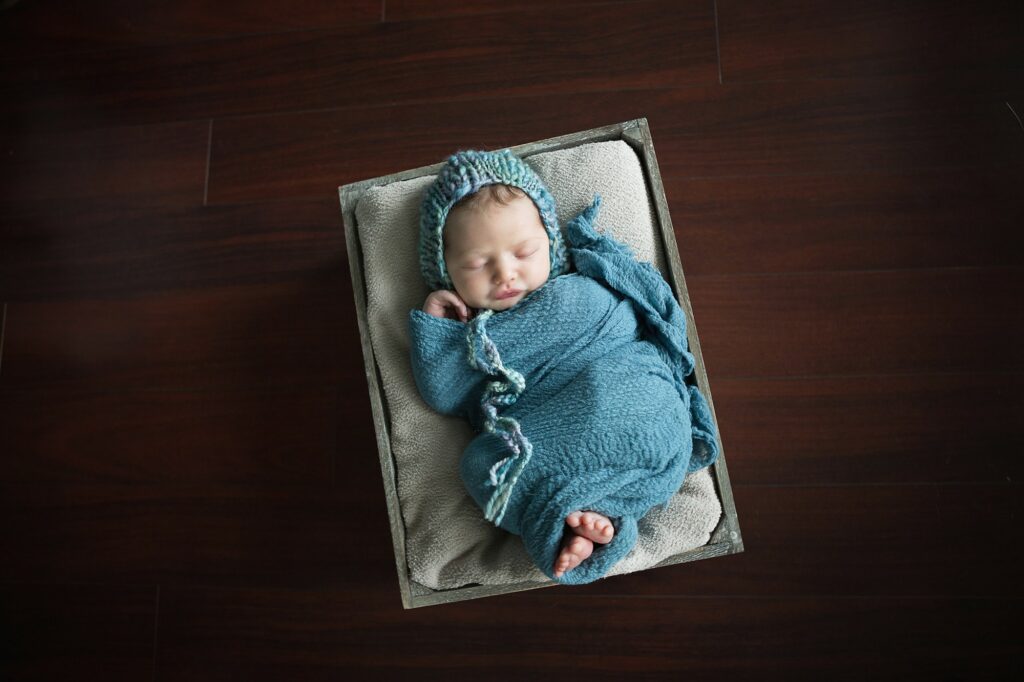 newborn in teal hat and wrap during photo session at home in a crate