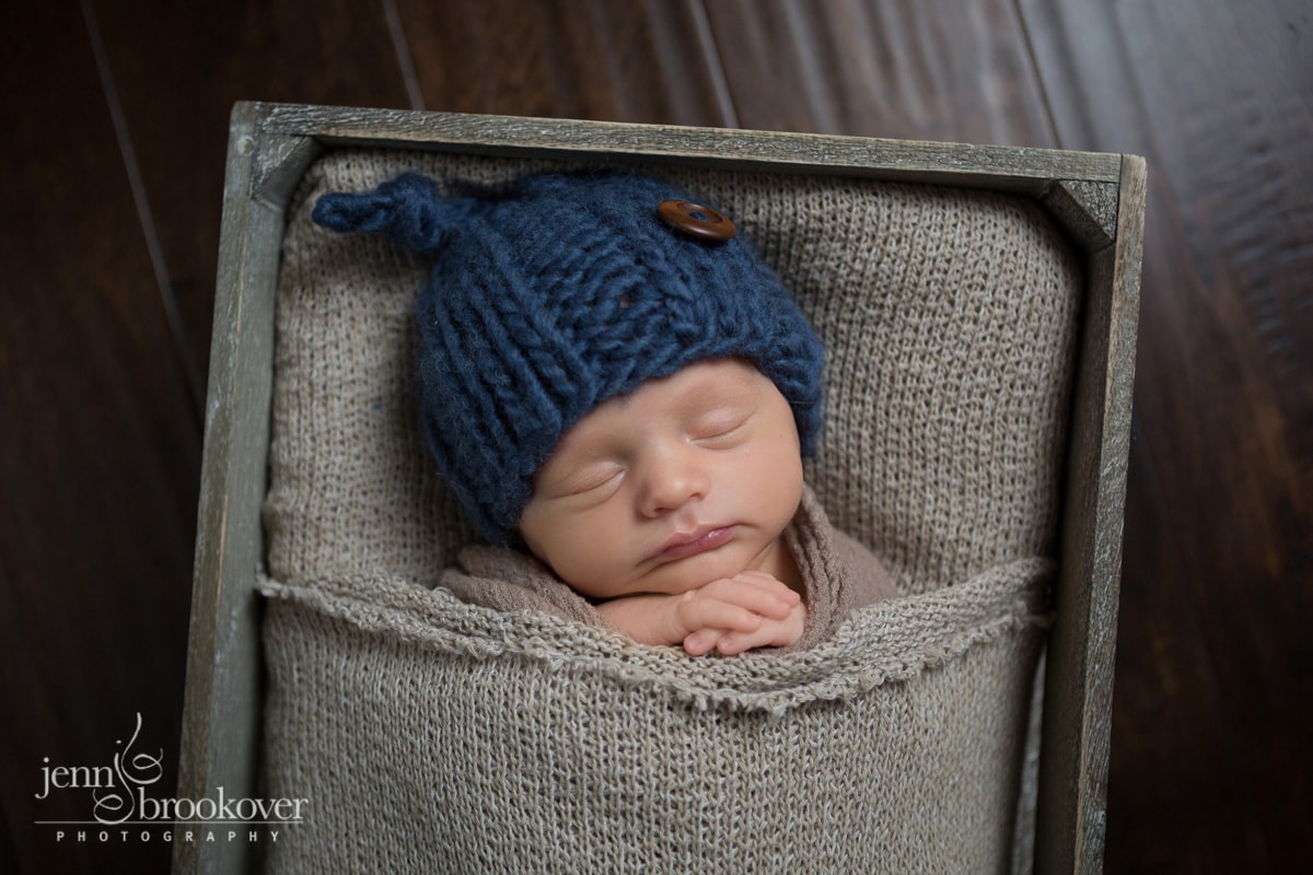 newborn boy in blue hat tucked into brown fabric bed on wood floor taken by Jenn Brookover