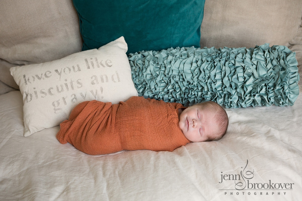 newborn wrapped in orange laying on a linen bed with pillows