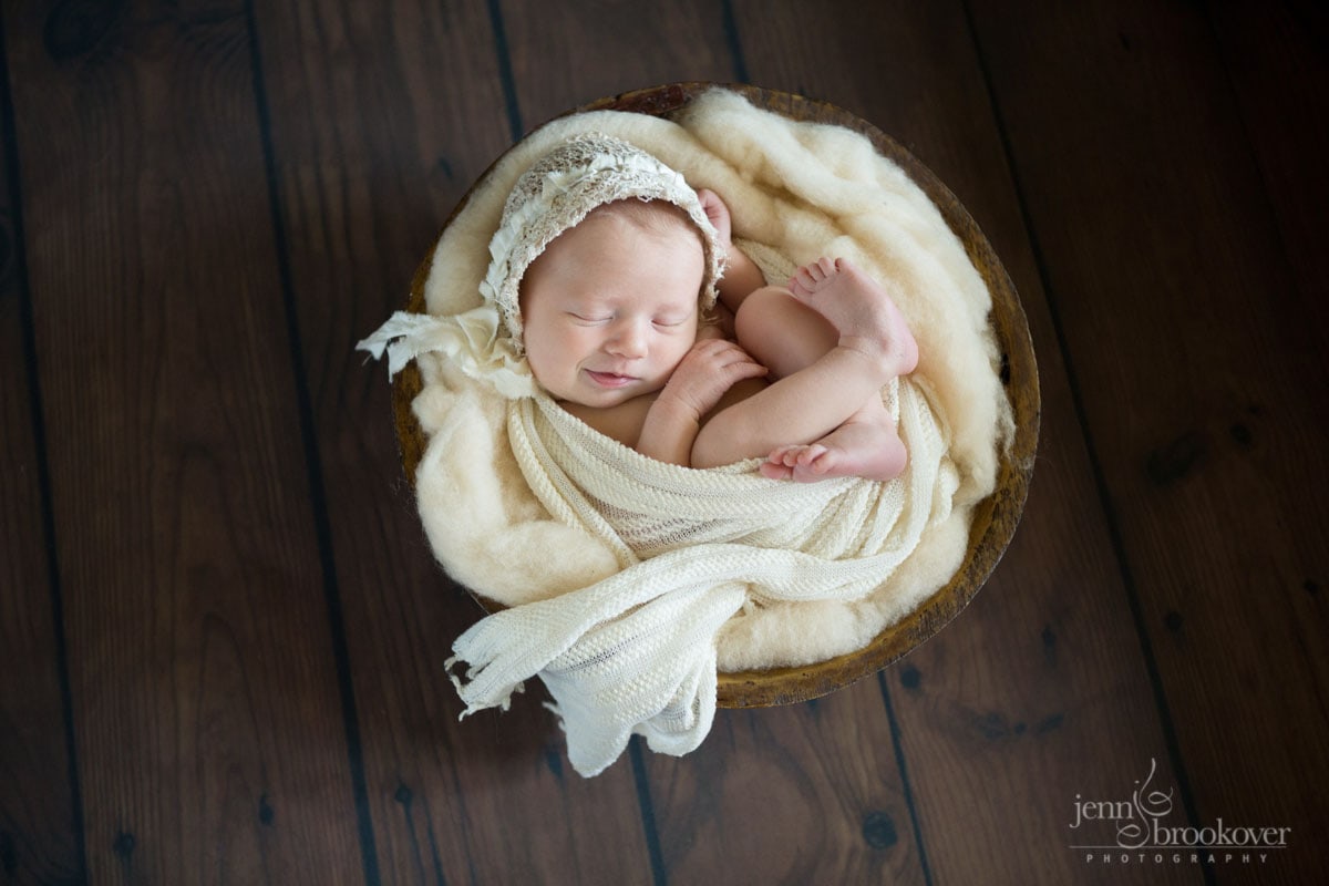 smiling baby wrapped in cream wool and fabric wearing a lace bonnet during photo session with Jenn Brookover
