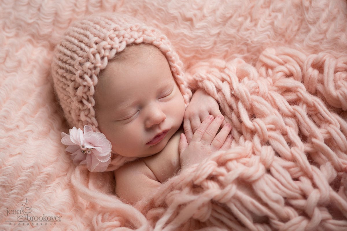 newborn baby girl in pink bonnet wth flower embellishment covered in knitted blanket taken by Jenn Brookover