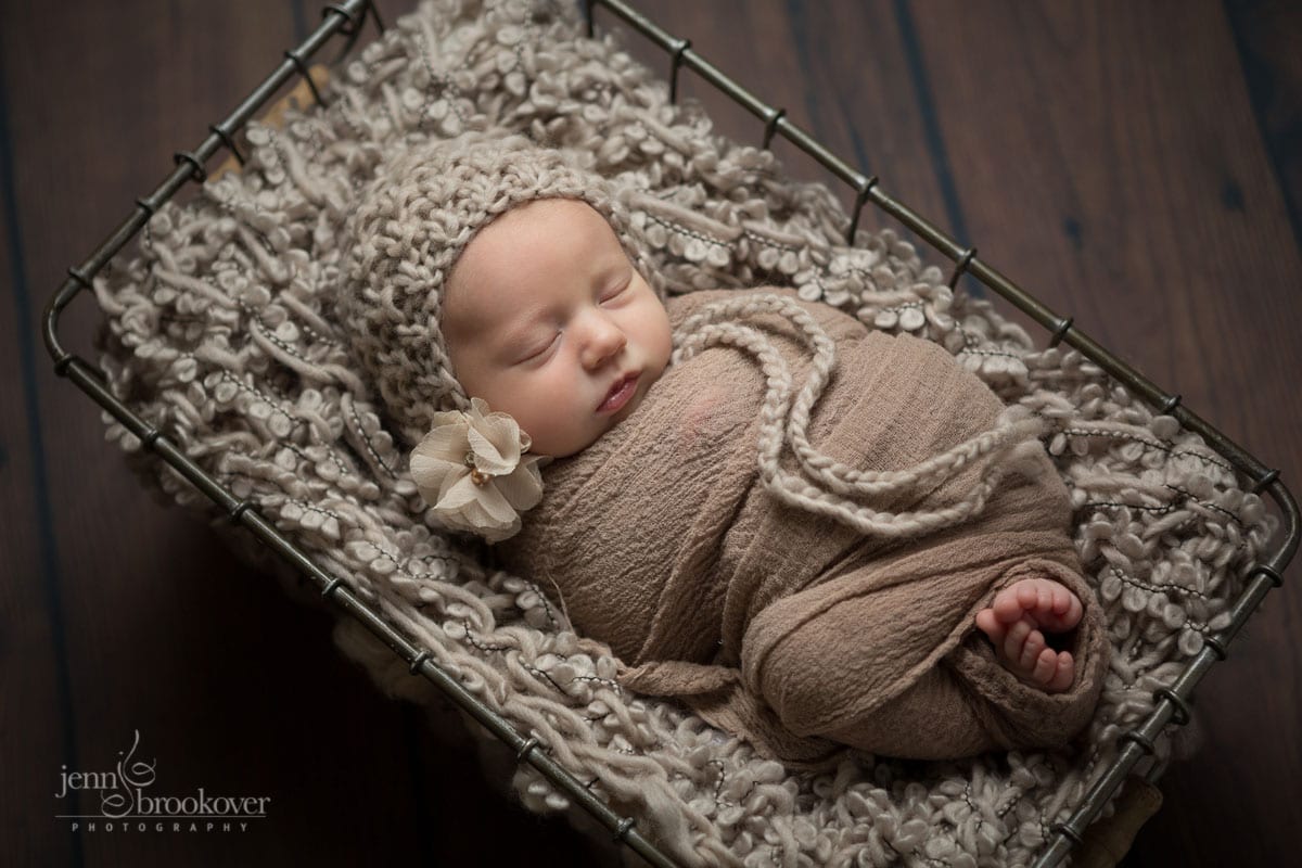 wrapped up two week old newborn girl in taupe wearing a bonnet with flower image by Jenn Brookover