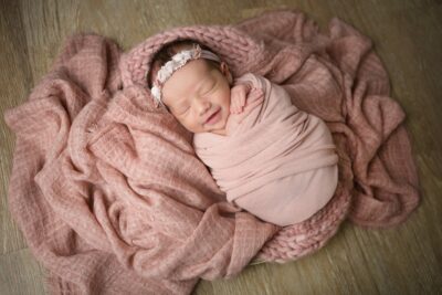 A newborn girl is wrapped in a pink blanket on a wooden floor.