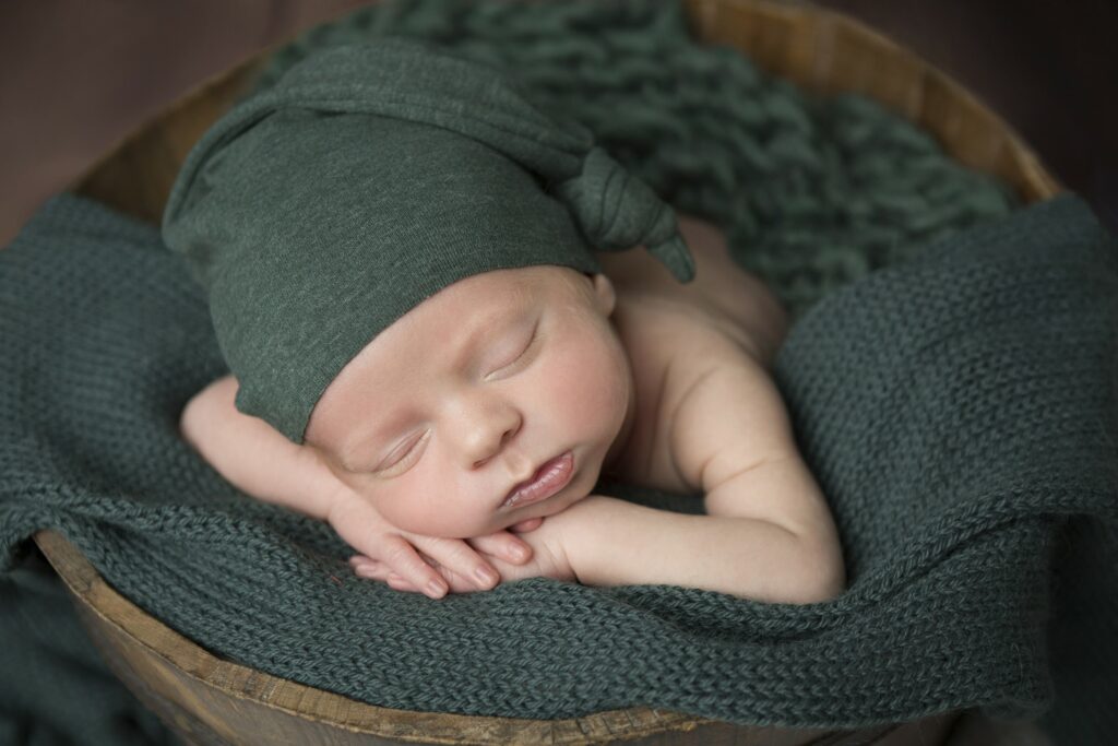 A baby boy in a green hat is sleeping in a wooden bowl.