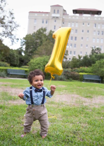 A baby boy holding a gold balloon in front of a building.