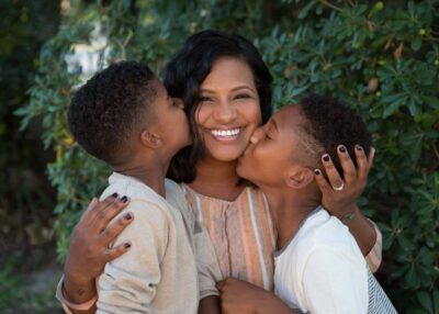 A woman kisses her sons in front of bushes.