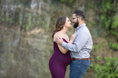 A pregnant couple embracing in front of a waterfall.