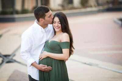 A pregnant couple kissing in front of a brick wall.