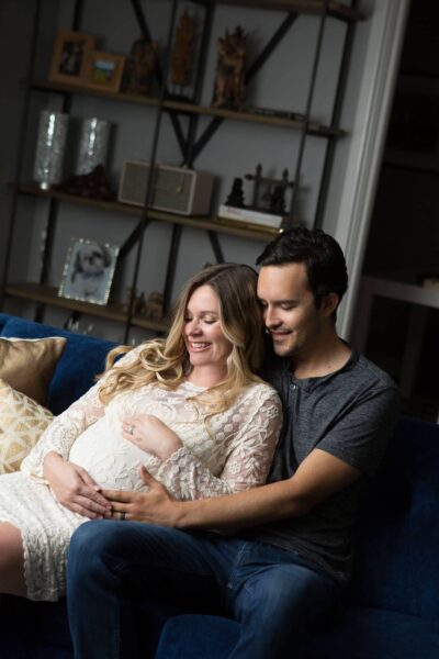 A pregnant couple sits on a couch in a living room.