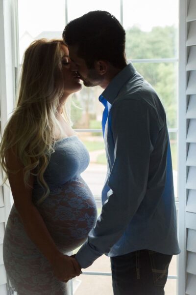 A pregnant couple kissing in front of a window.