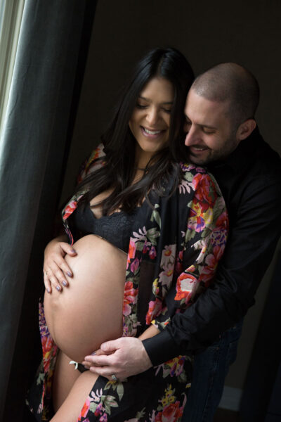 A pregnant woman is posing with her husband in front of a window.