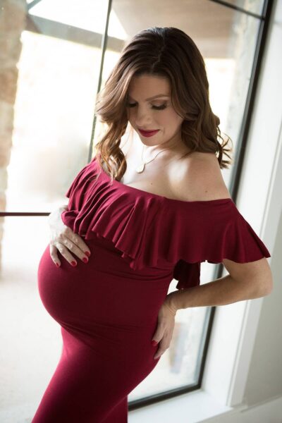 A pregnant woman in a burgundy dress posing in front of a window.