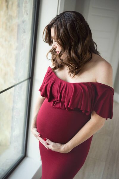 A pregnant woman in a burgundy dress is standing in front of a window.