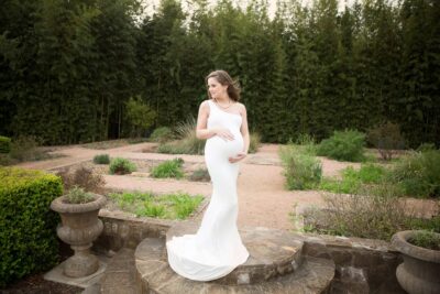A pregnant woman in a white dress standing on steps in a garden.