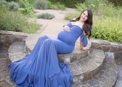 A pregnant woman in a blue dress sits on steps in a garden.