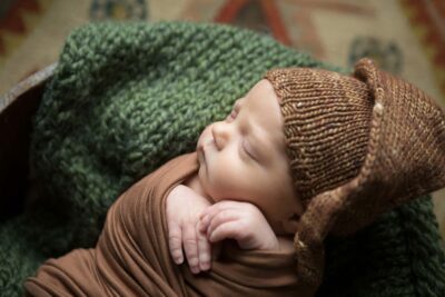 A baby wearing a brown knitted hat is laying in a basket.