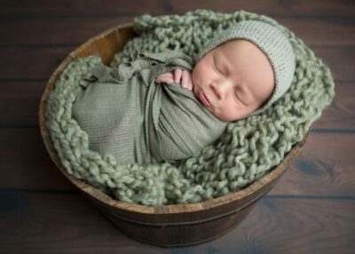 A newborn sleeping in a green knitted hat in a wooden bucket.