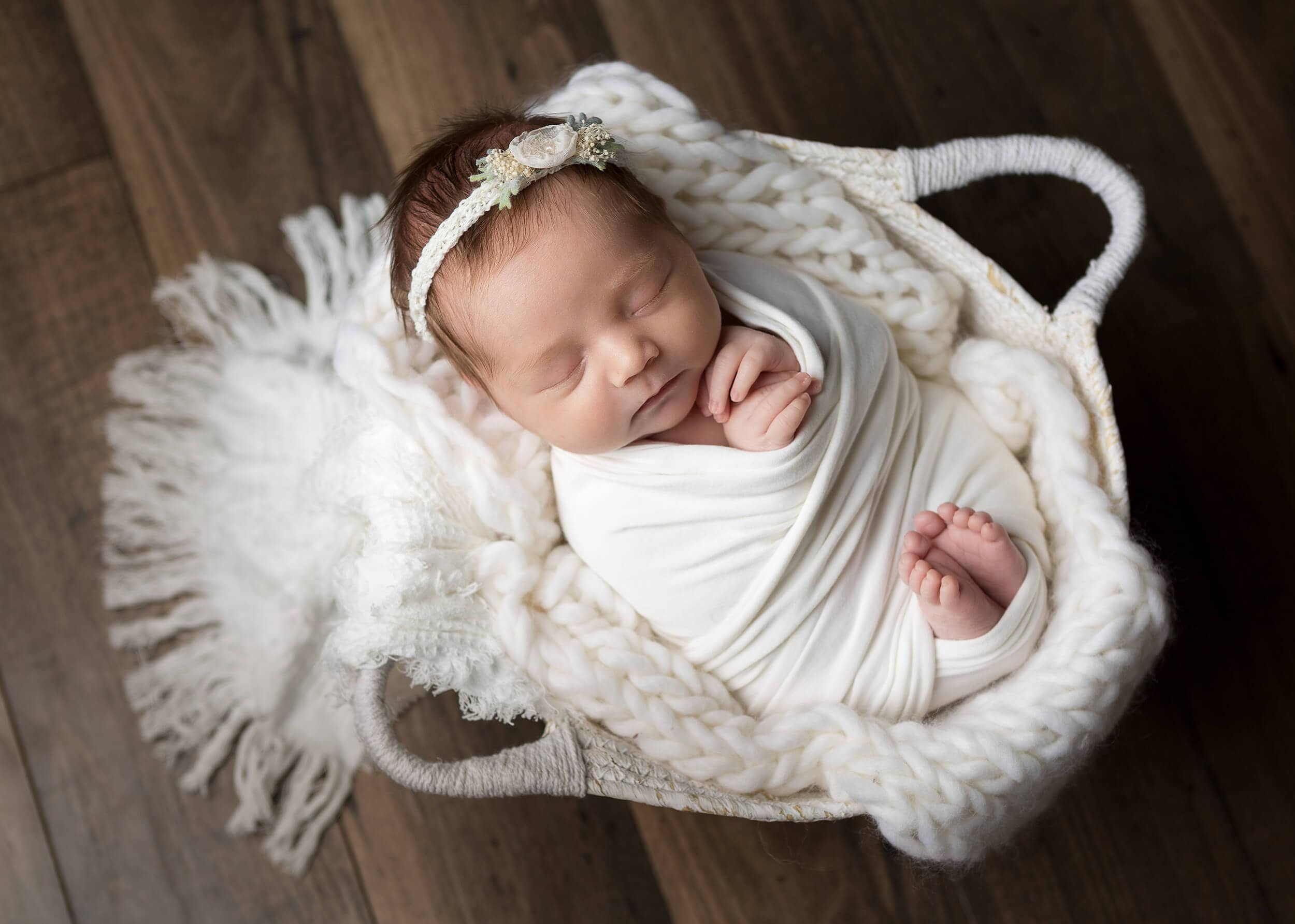A newborn girl is wrapped in a white basket on a wooden floor.