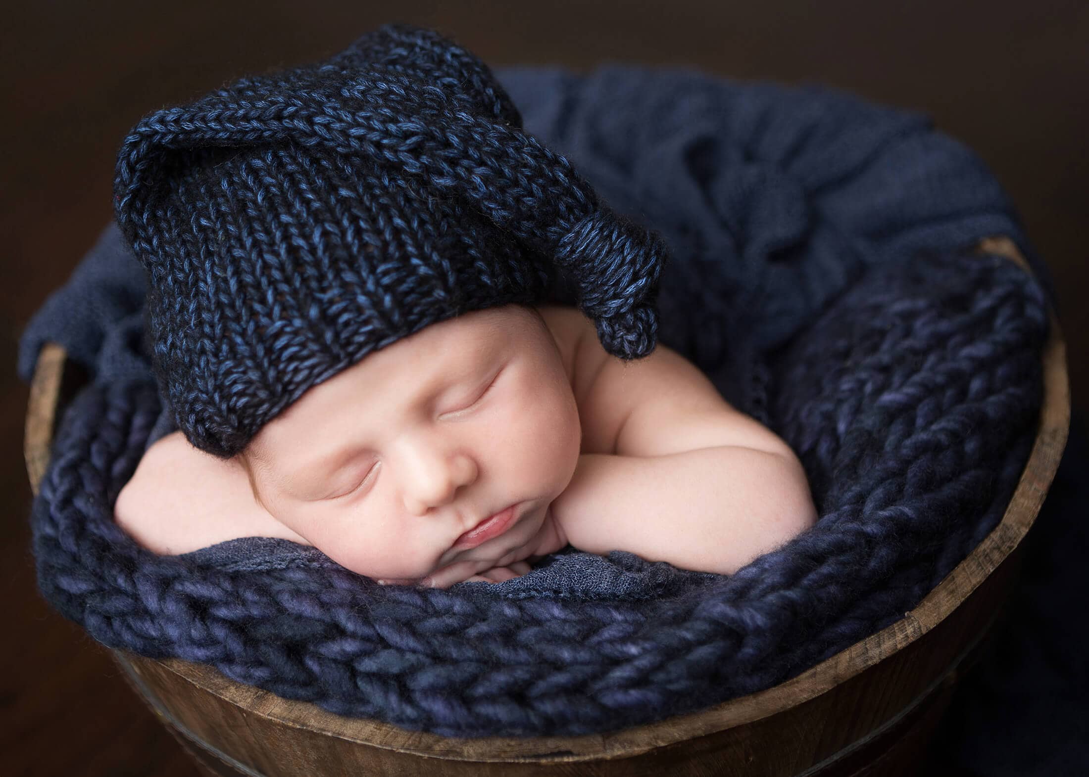 A newborn sleeping in a blue knitted hat in a wooden bowl.