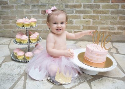 A baby girl in a tutu sitting next to a cake and cupcakes.