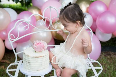 A baby girl sitting on a bench with balloons and a cake.