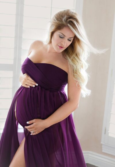 A pregnant woman in a purple dress posing in front of a window.