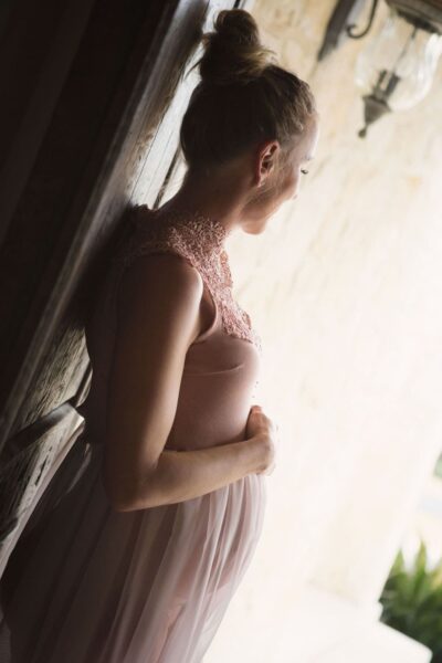 A pregnant woman in a pink dress leaning against a door.