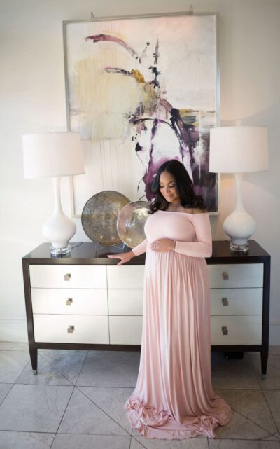 A pregnant woman in a pink dress standing in front of a dresser.