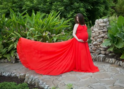 A pregnant woman in a red dress posing in a garden.