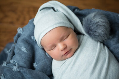 A newborn sleeping in a blue blanket with a grey pom pom.
