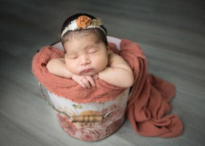 A newborn girl sleeping in a bucket on a wooden floor.