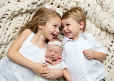 Three children laughing while laying on a blanket with a newborn baby.
