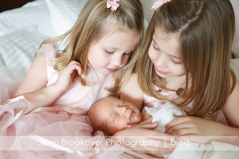 Three little girls in pink dresses holding a newborn baby captured by Jenn Brookover Photography.