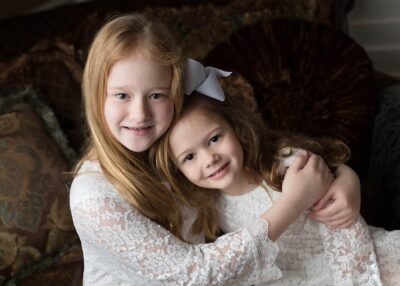 Two little girls in white dresses hugging on a couch.