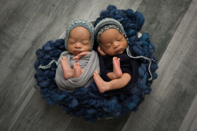 Two newborn twins in blue hats laying on a wooden floor.