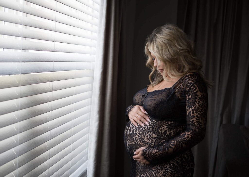 A pregnant woman in a black lace dress standing by a window.